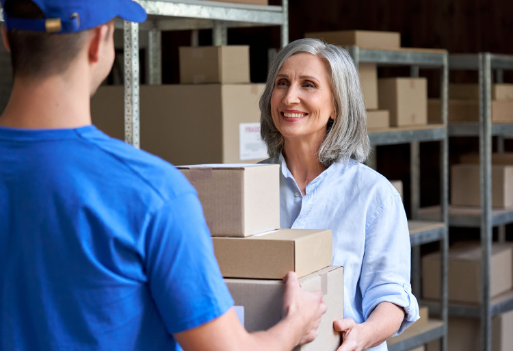 Older woman in a warehouse handing a stack of boxes to a man